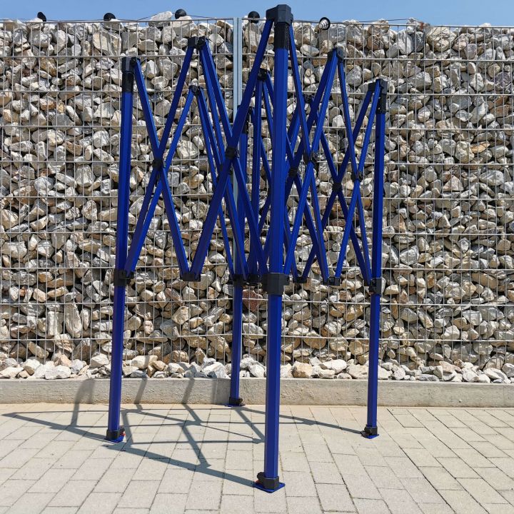 Uppdelad, men oskjuten blå konstruktion (ram) av en saxpartytält, stående på plattor med småstenar i bakgrunden (vid staketet).